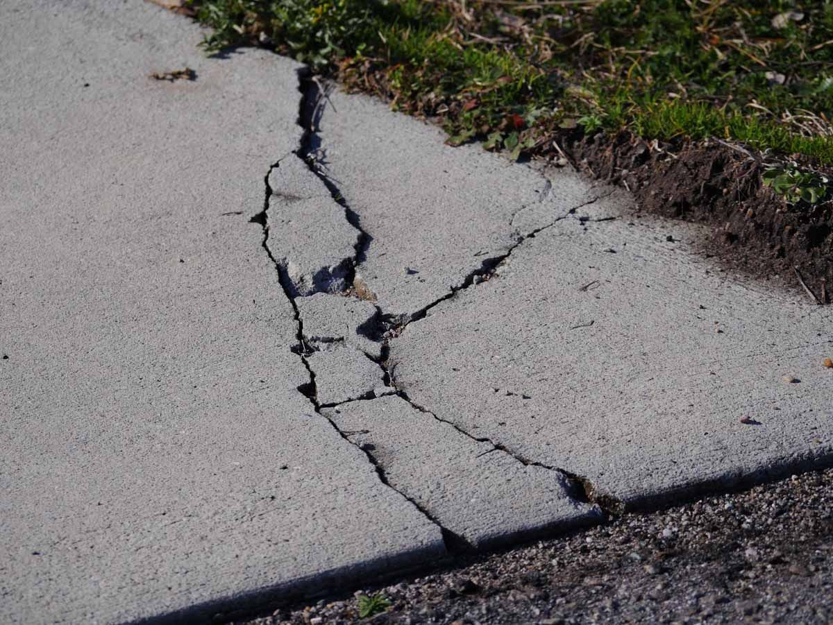 A large, deep crack splits a grey concrete sidewalk near a grassy edge.