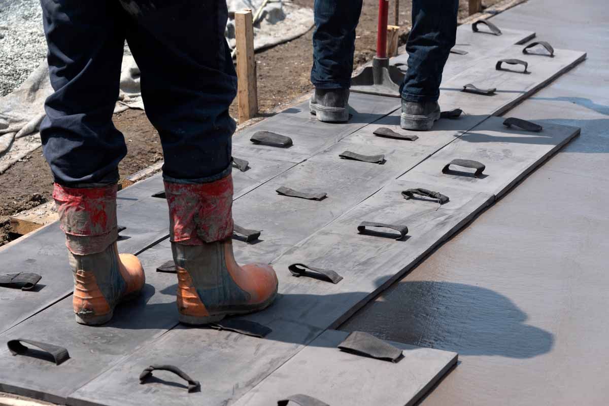 Construction workers standing on black rubber concrete mats laid over a freshly poured wet concrete sidewalk.