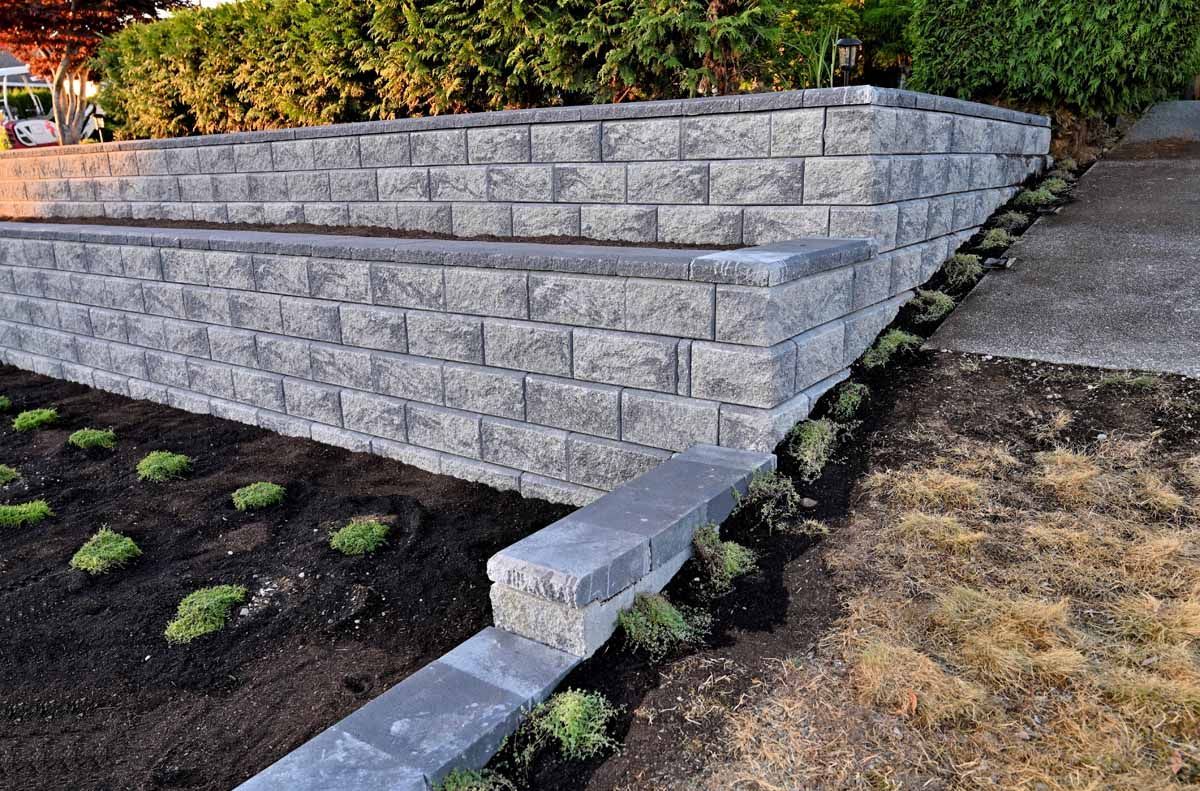 A two-tiered gray concrete block retaining wall structure next to a garden bed and a stone path.