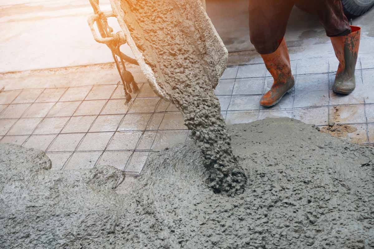 Wet cement pours from a truck chute onto a paved surface, with a worker in rubber boots standing nearby.