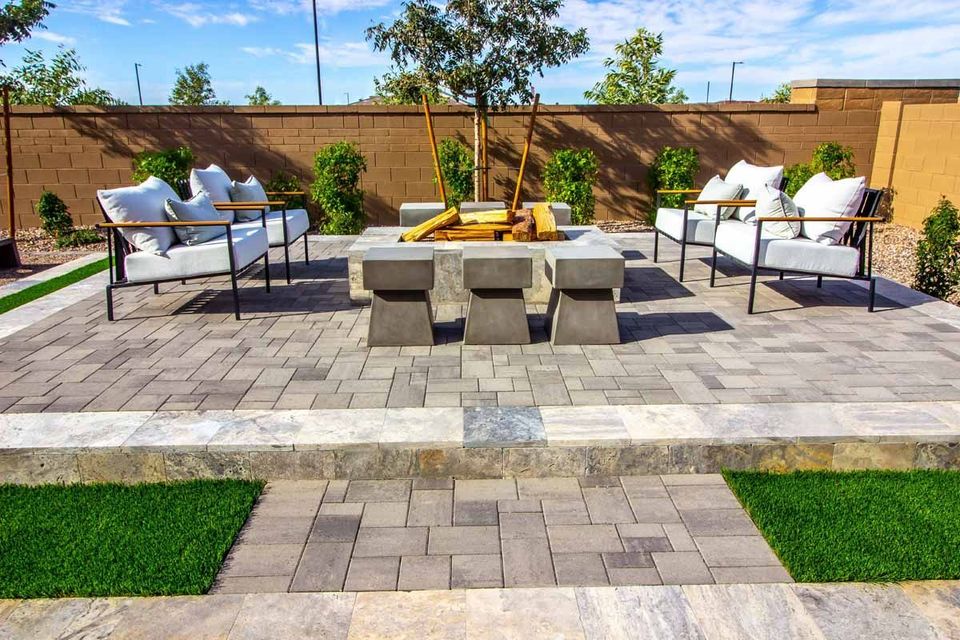 A patio with stone pavers, a central fire pit, two sets of white outdoor armchairs, and surrounding green turf.