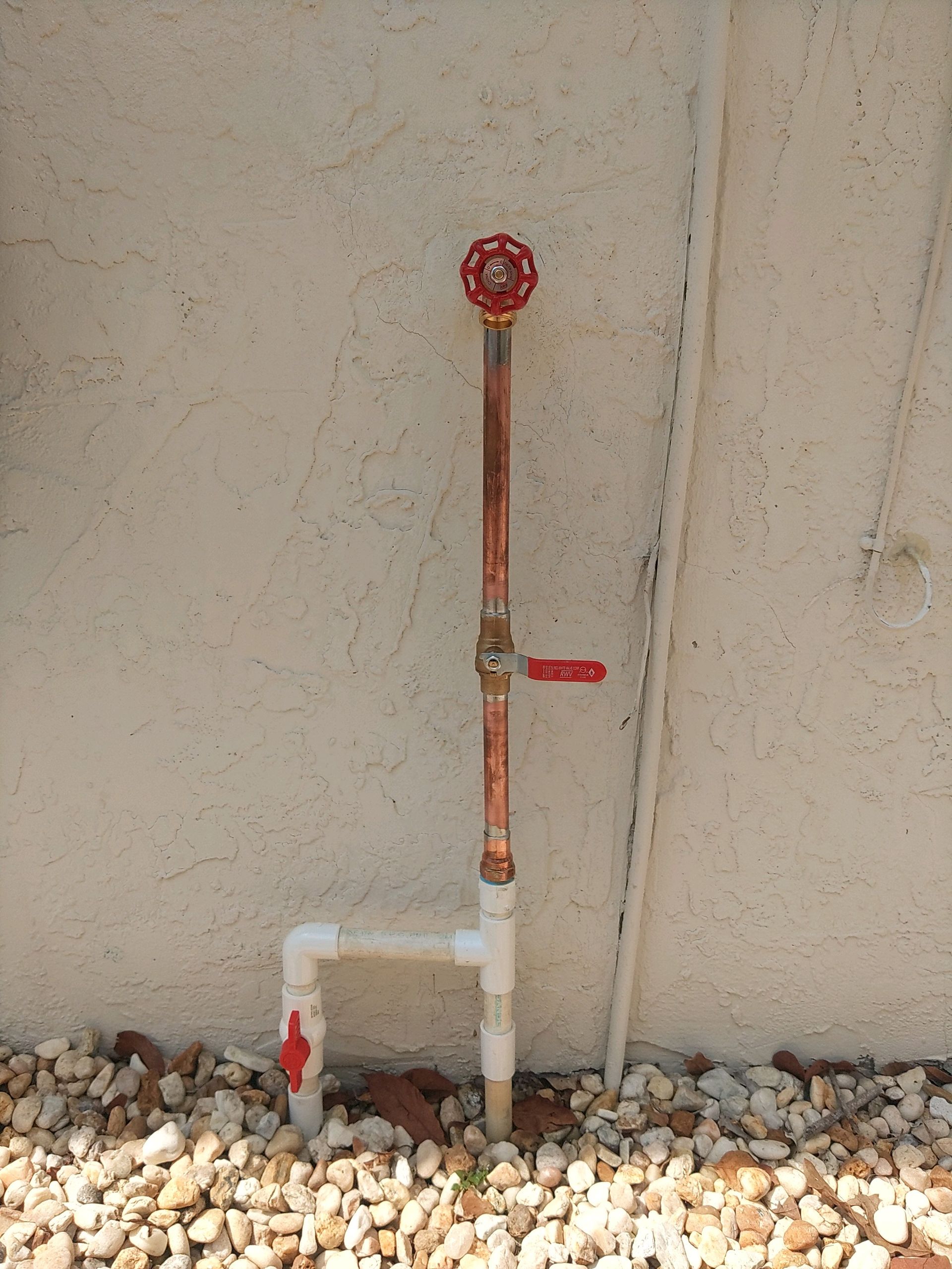 Outdoor plumbing setup with PVC and copper pipes, featuring multiple valves against a stucco wall over gravel.