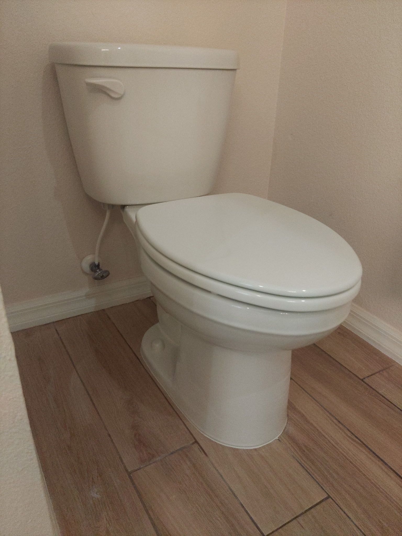 A white toilet with a closed lid sits in a corner against neutral walls on a wood-look tile floor.
