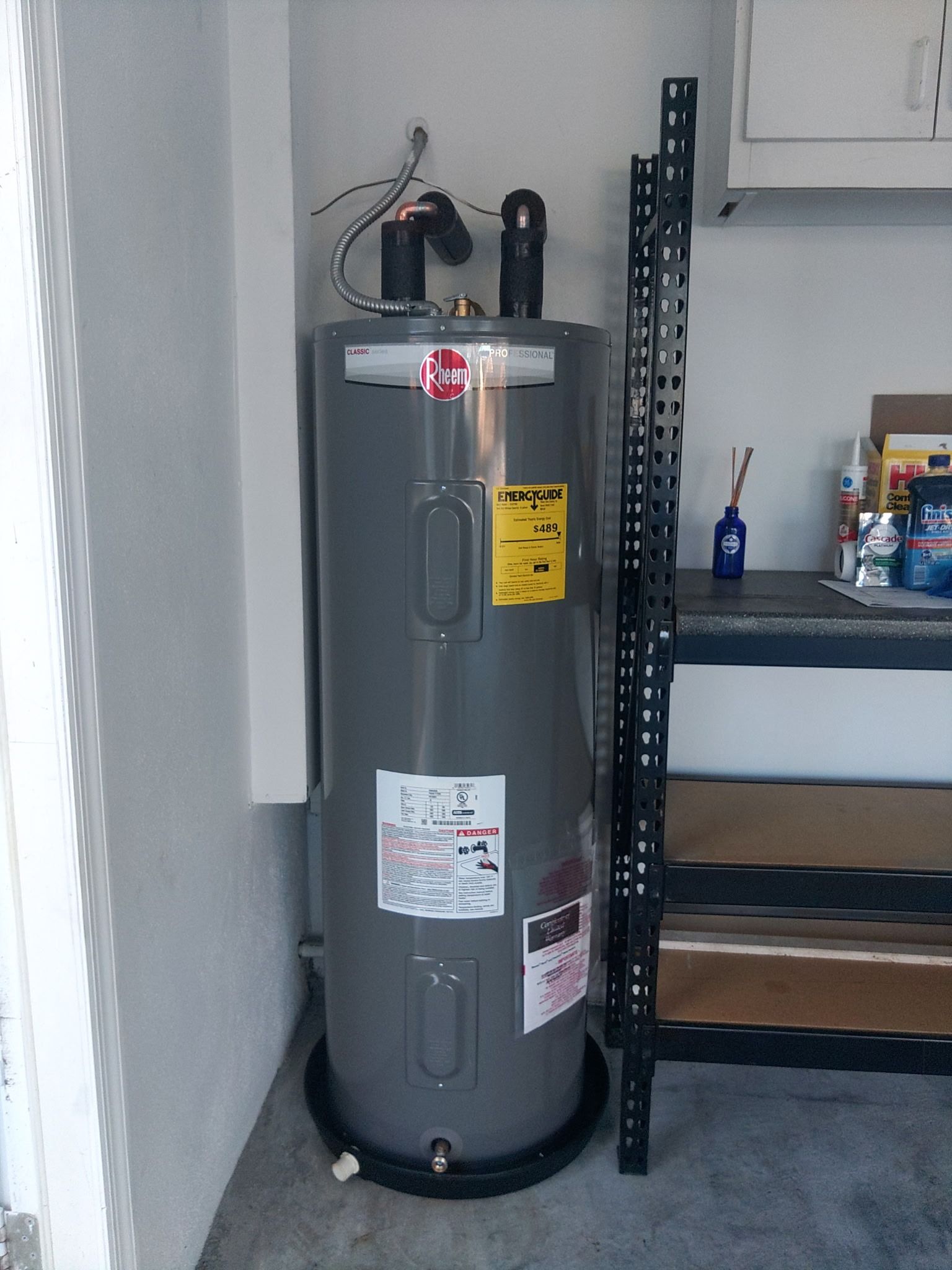 A gray residential water heater sits in a corner inside a garage next to a metal shelving unit.