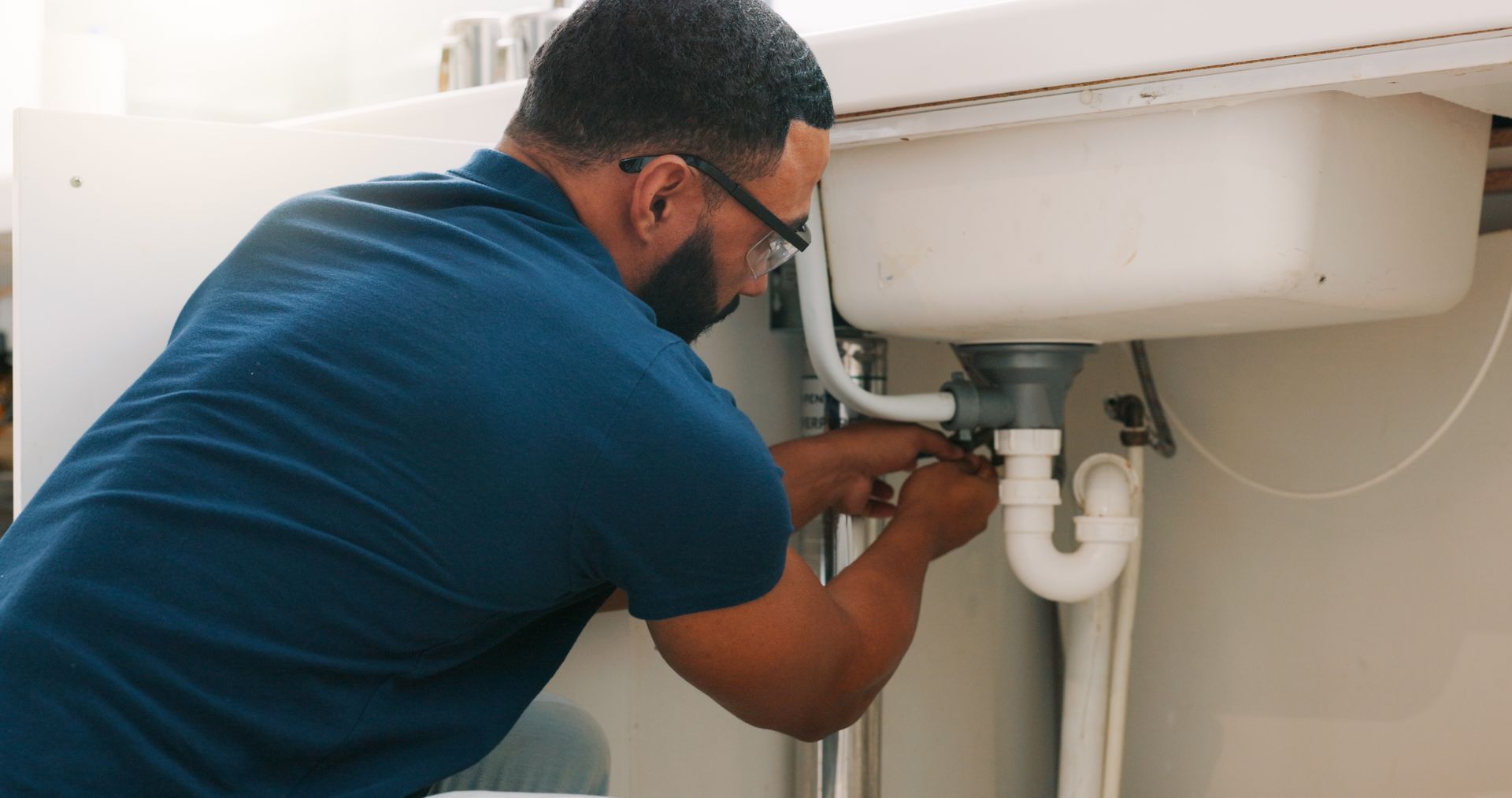 A plumber tightening a white PVC drain pipe beneath a kitchen sink using a wrench
