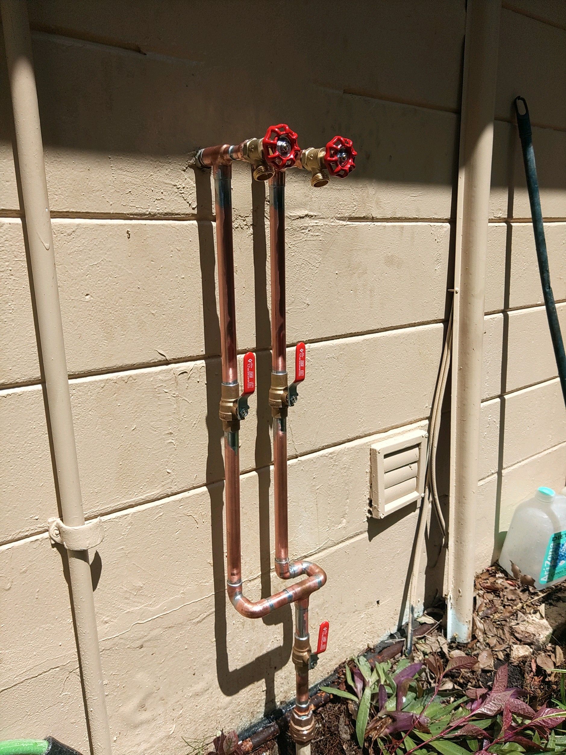 Copper pipes with red-handled valves and faucets installed vertically against a tan cinder block wall.