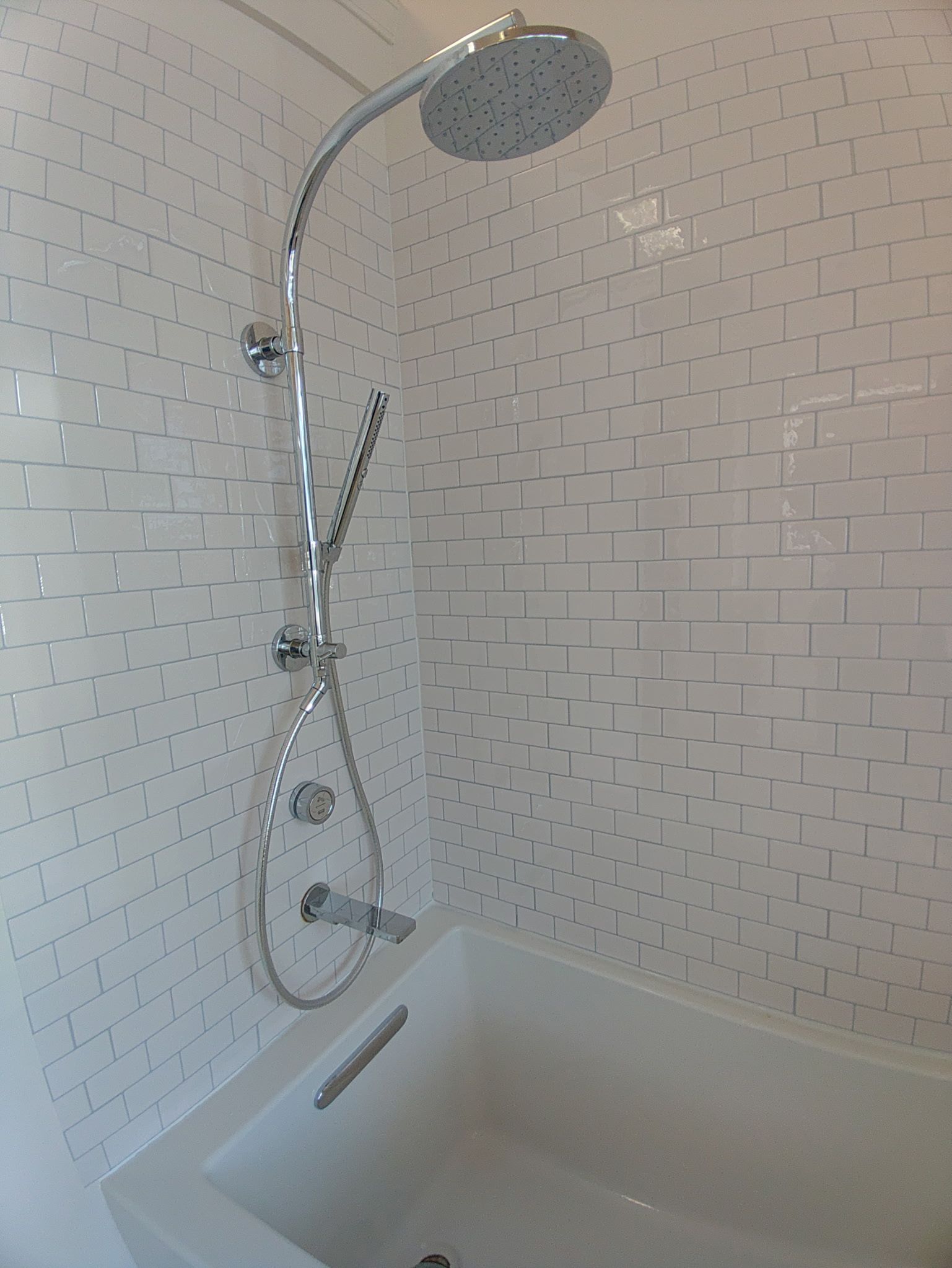 A chrome shower head and handle mounted on a wall with white, vertical, basket-weave patterned tiles above a bathtub.