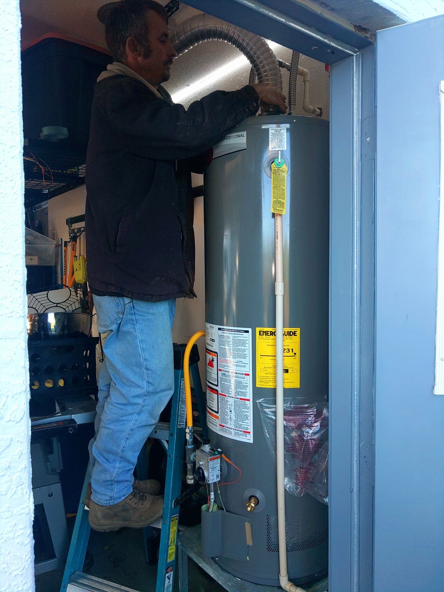 A person stands on a step ladder to work on a tall, gray water heater inside a utility closet.