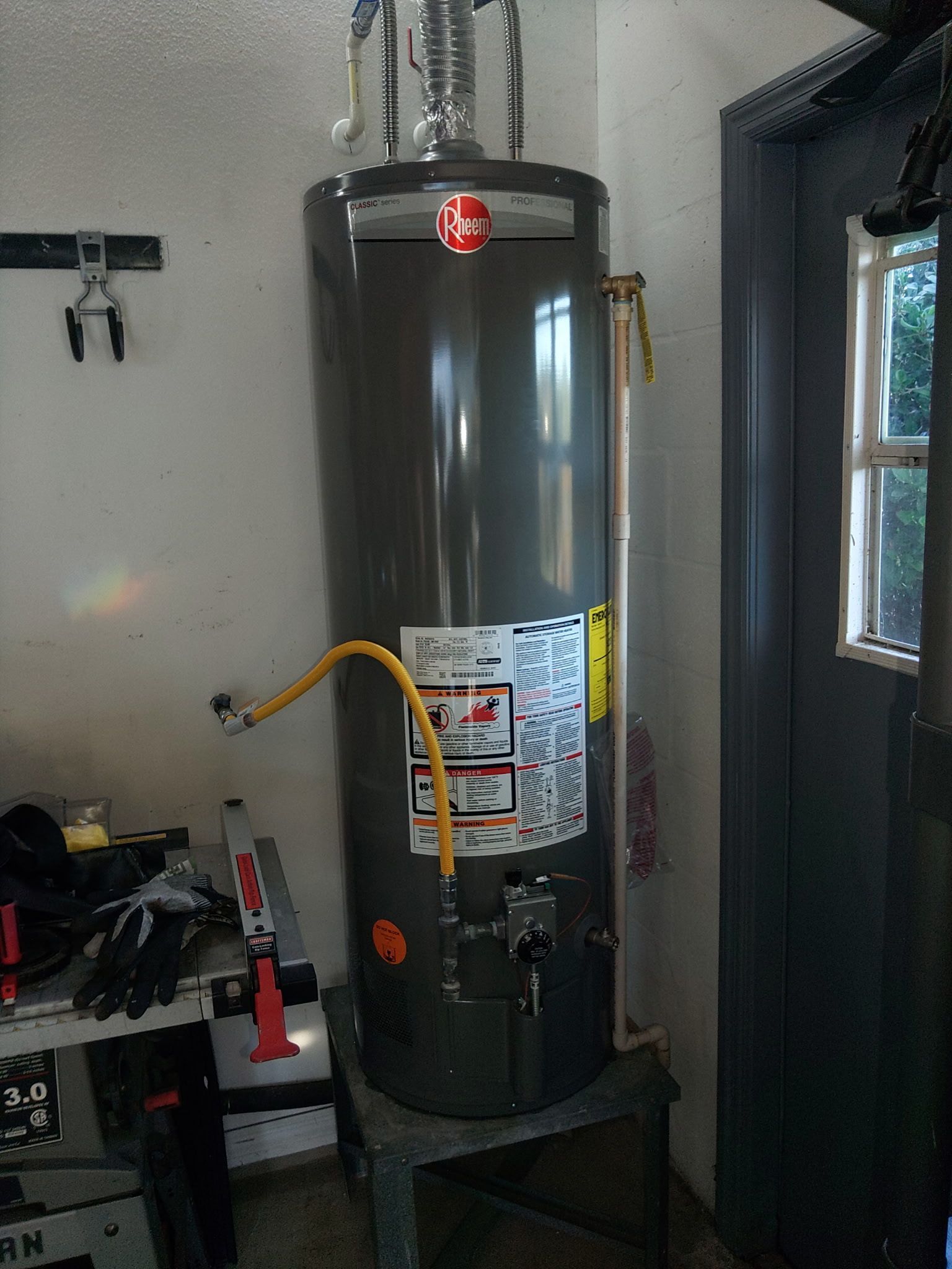 A grey Rheem gas water heater standing on a metal platform in a garage near a doorway.