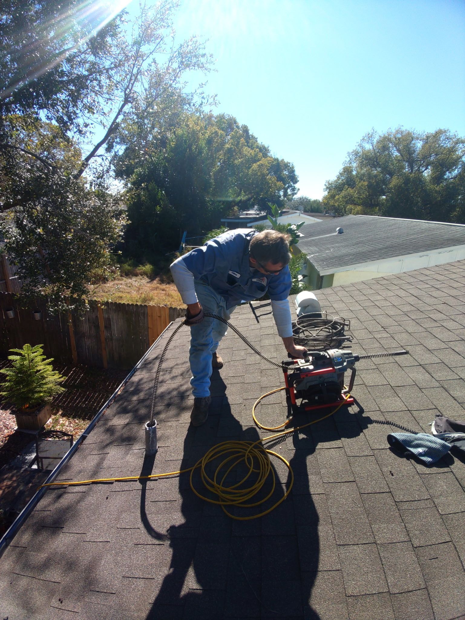 A person in a blue shirt works on a residential roof, operating machinery with cables spread across the shingles.