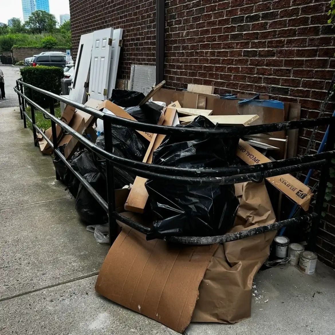 Overflowing cardboard boxes and trash bags piled behind a black metal barrier on a sidewalk by a brick wall