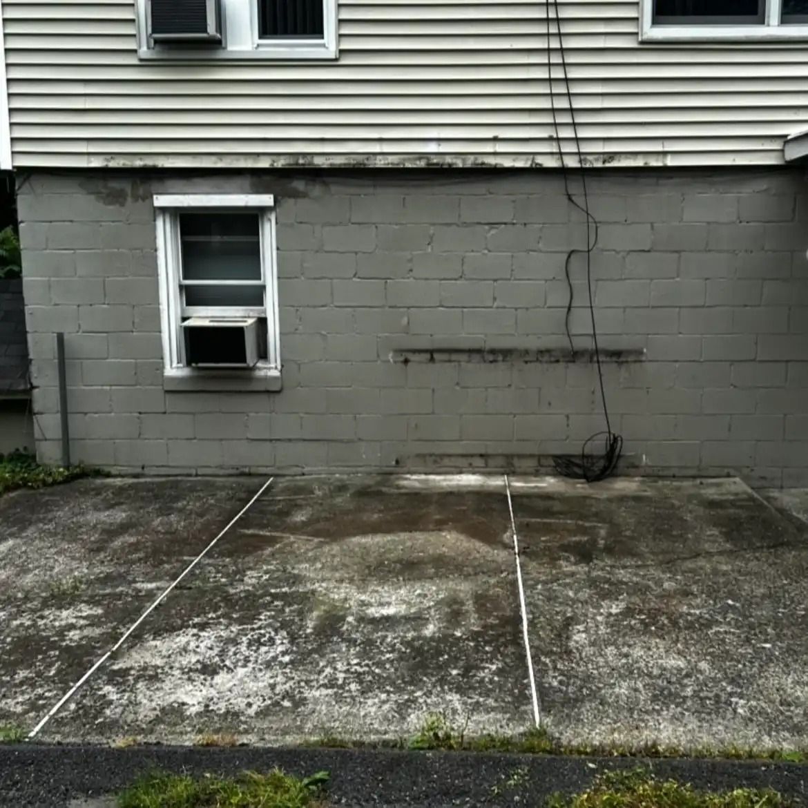 Cracked gray concrete wall of a house with two basement windows and a worn patio slab in front