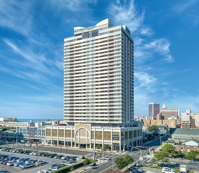 Tall white high-rise tower by the waterfront under a blue sky, with a parking lot in front.