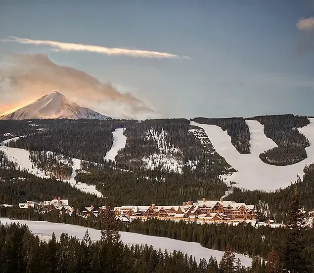 Snowy mountain ski resort with groomed slopes, forest, and chalets under a cloudy sunset sky
