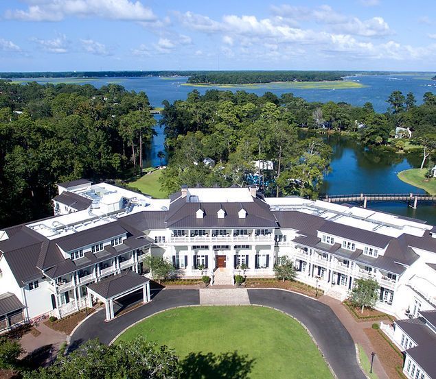 Aerial view of a white waterfront estate with a circular driveway, surrounded by trees and blue water beyond.