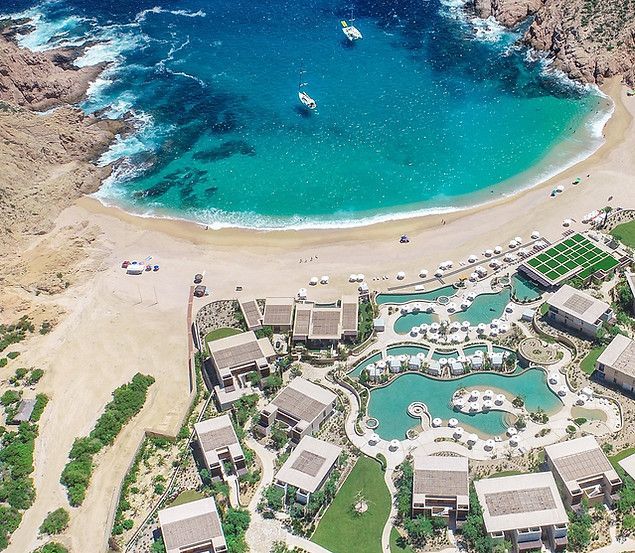 Aerial view of a turquoise cove beach beside a resort with pools and tan buildings