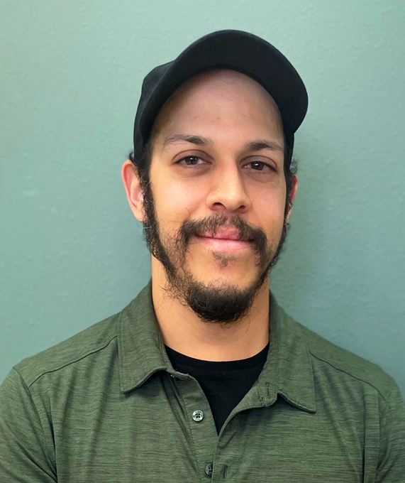 Man with dark hair and beard, wearing a black cap and green button-down shirt, smiles in front of a teal wall.