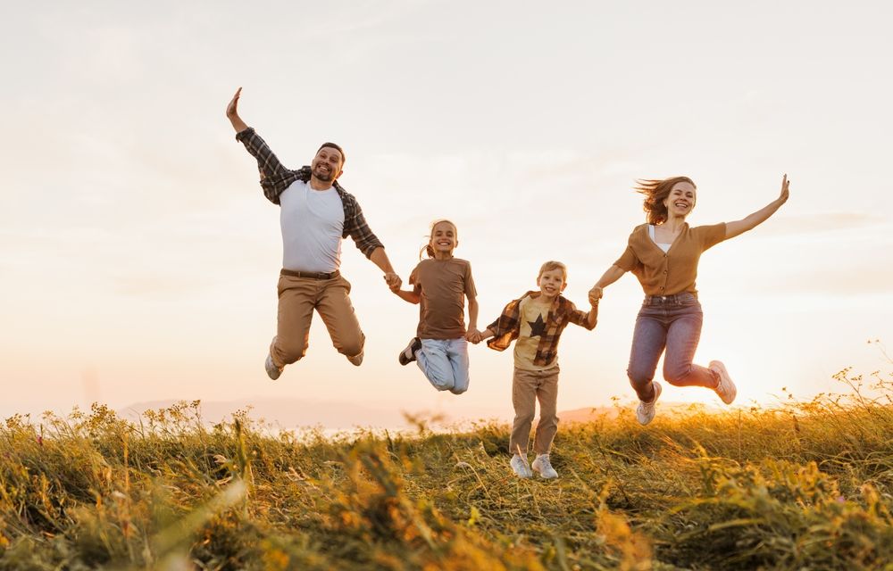 Family of four jumps in joy on a grassy hill at sunset, holding hands and smiling.
