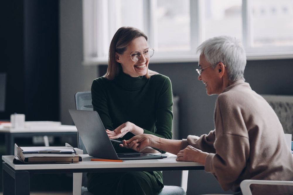 Woman and older person at a desk looking at a laptop, smiling. Indoor office with window.