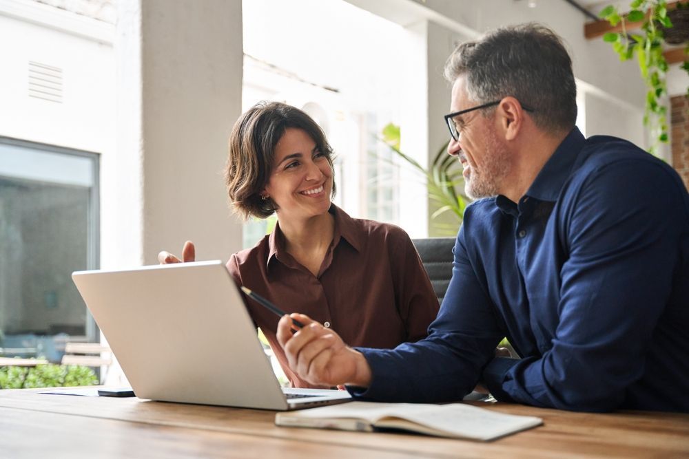 Two people looking at laptop, smiling, discussing something at a desk indoors.