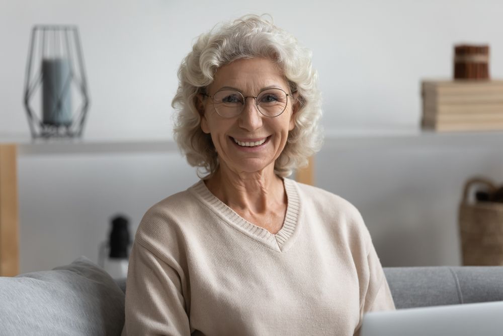Smiling woman with glasses in a beige sweater, sitting indoors.