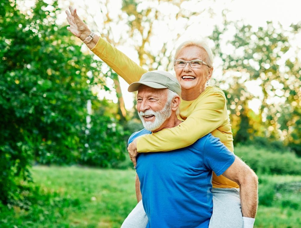Man giving woman a piggyback ride outdoors; both smiling, green trees in the background.