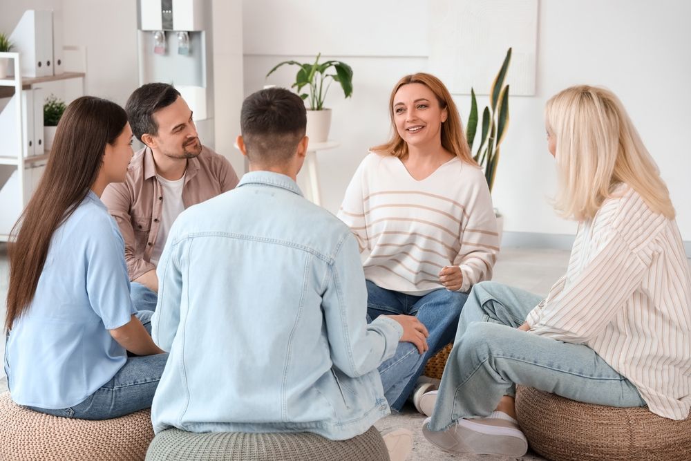 People in a circle, one woman speaking, others listening attentively. Indoor setting, light colors.