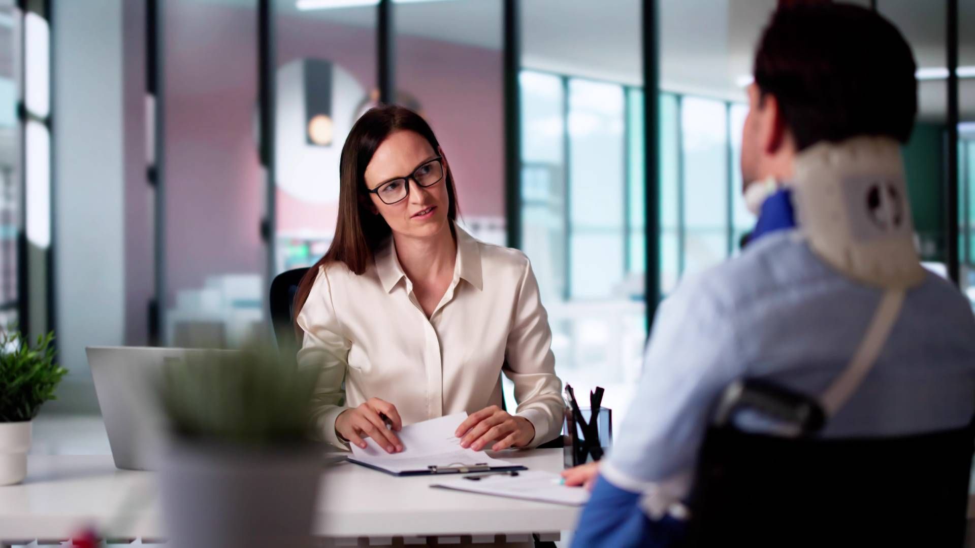 Woman in glasses interviewing a person wearing a neck brace, seated at a desk in an office.