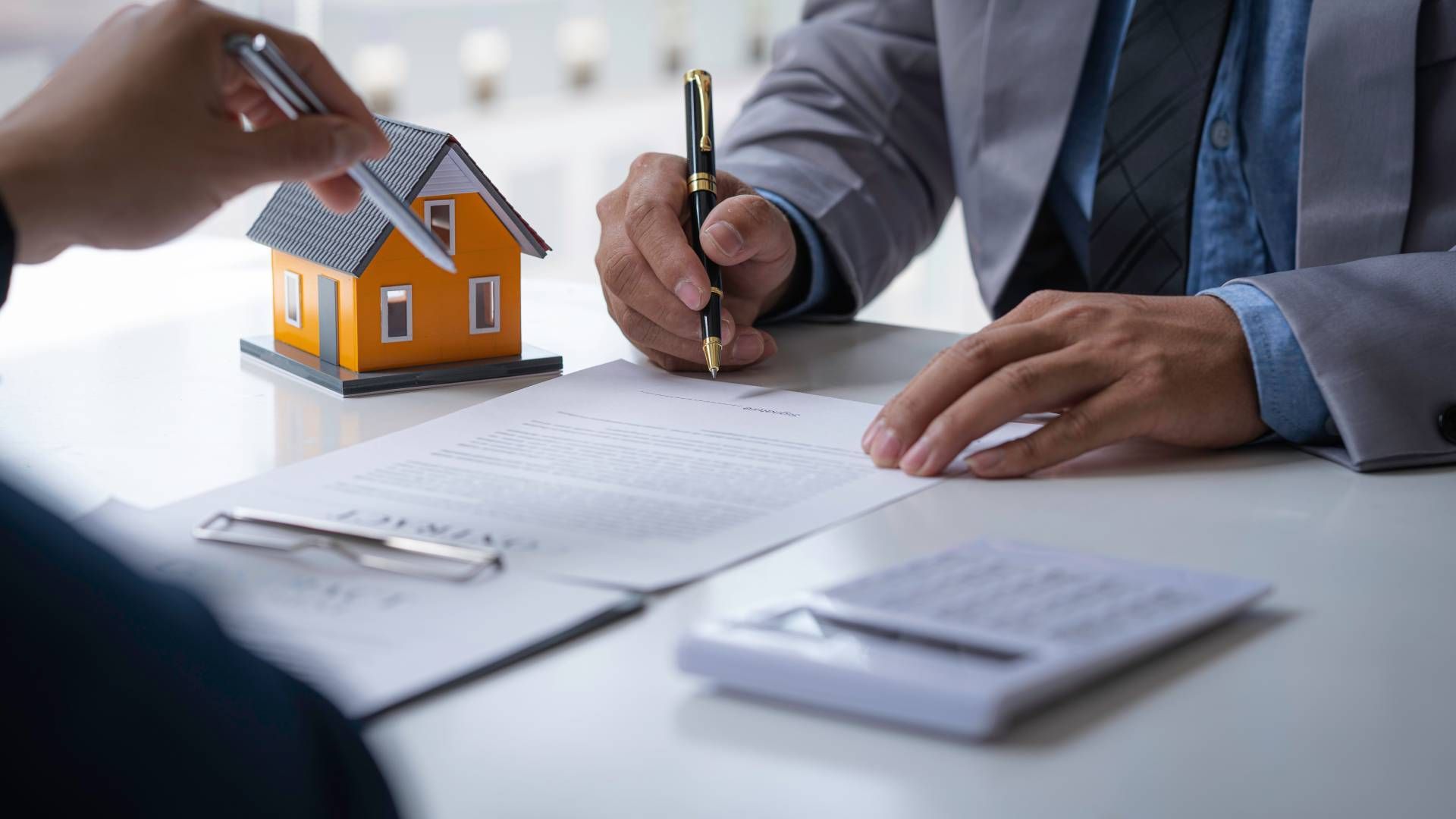 Two people at a desk, one signing a contract. A model house and calculator are present.