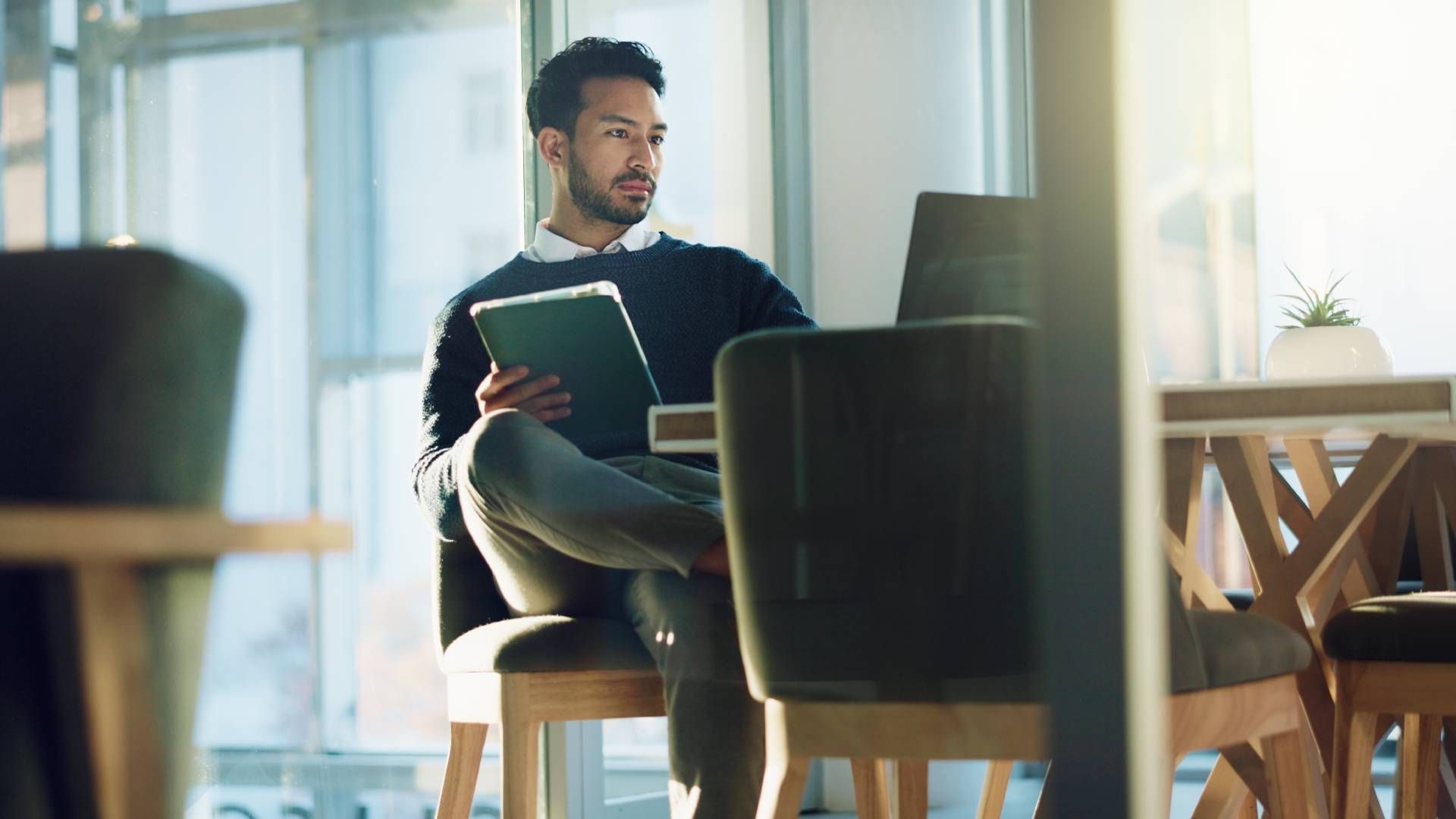 Man sitting in a chair, holding a tablet, looking away from a laptop at a table by a window.