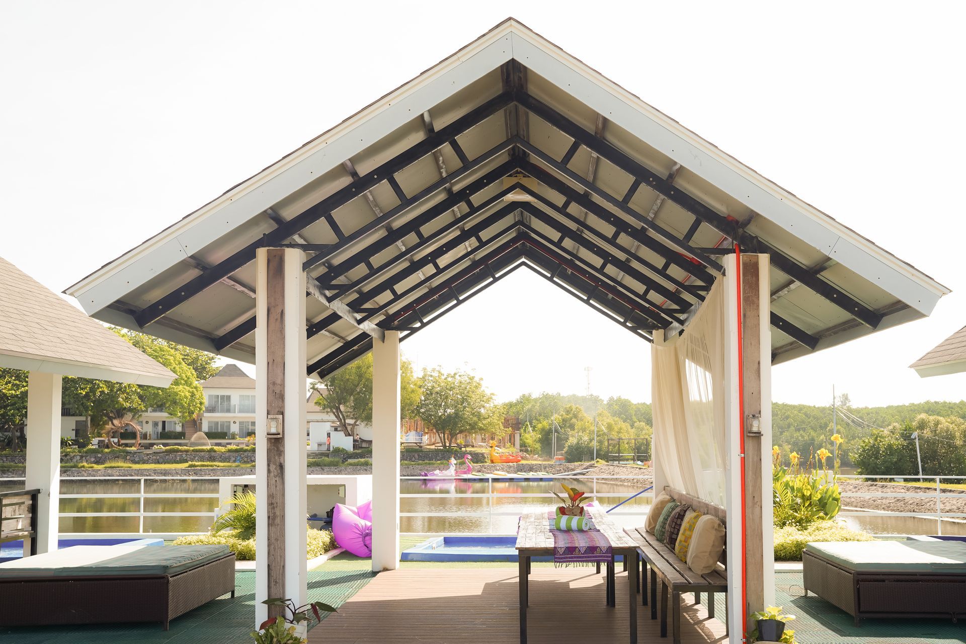 Gazebo over water with table, chairs, cushions. Trees and buildings in the background. Sunny day.