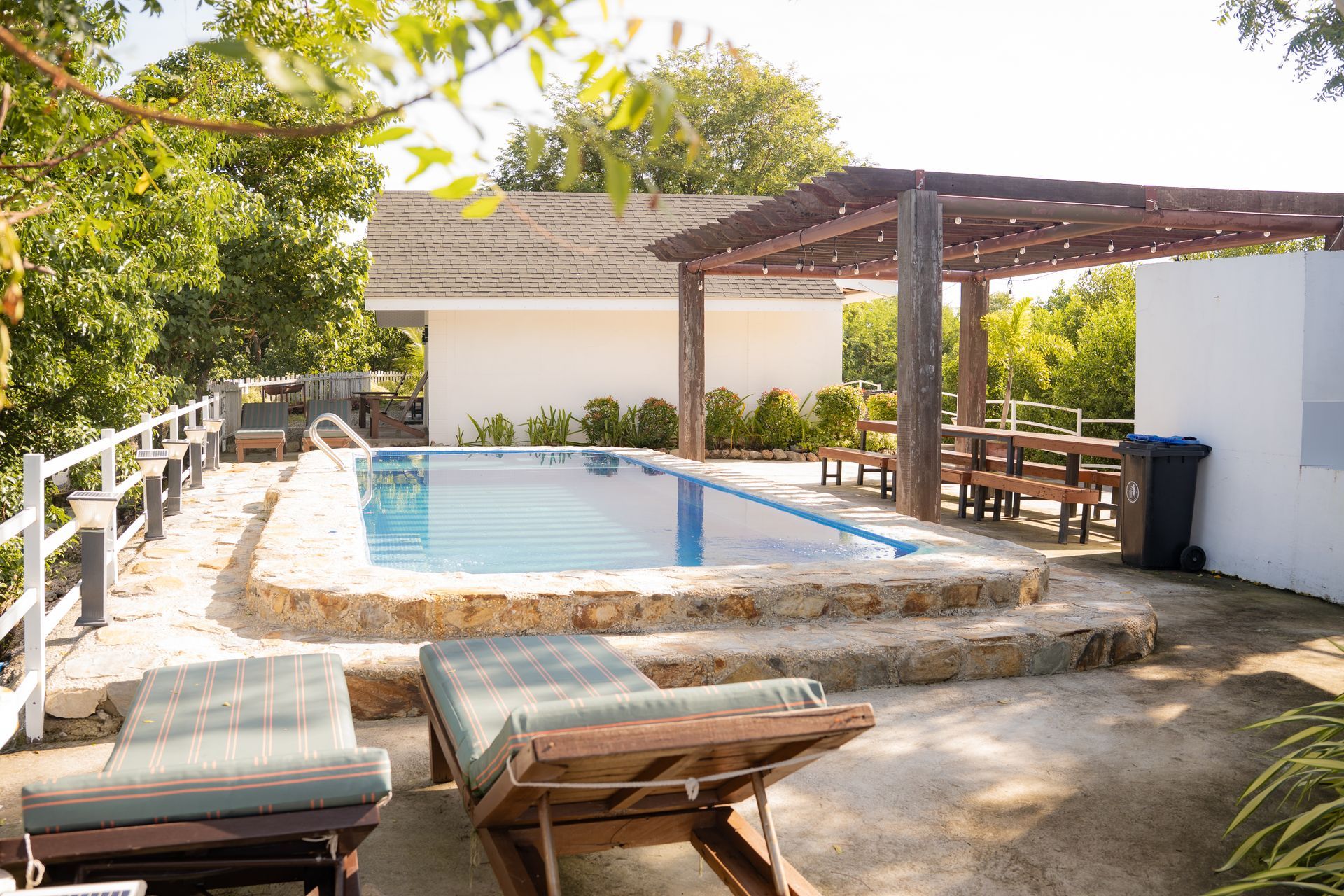 Pool with stone surround, sun loungers, pergola with seating, white building, and trees.