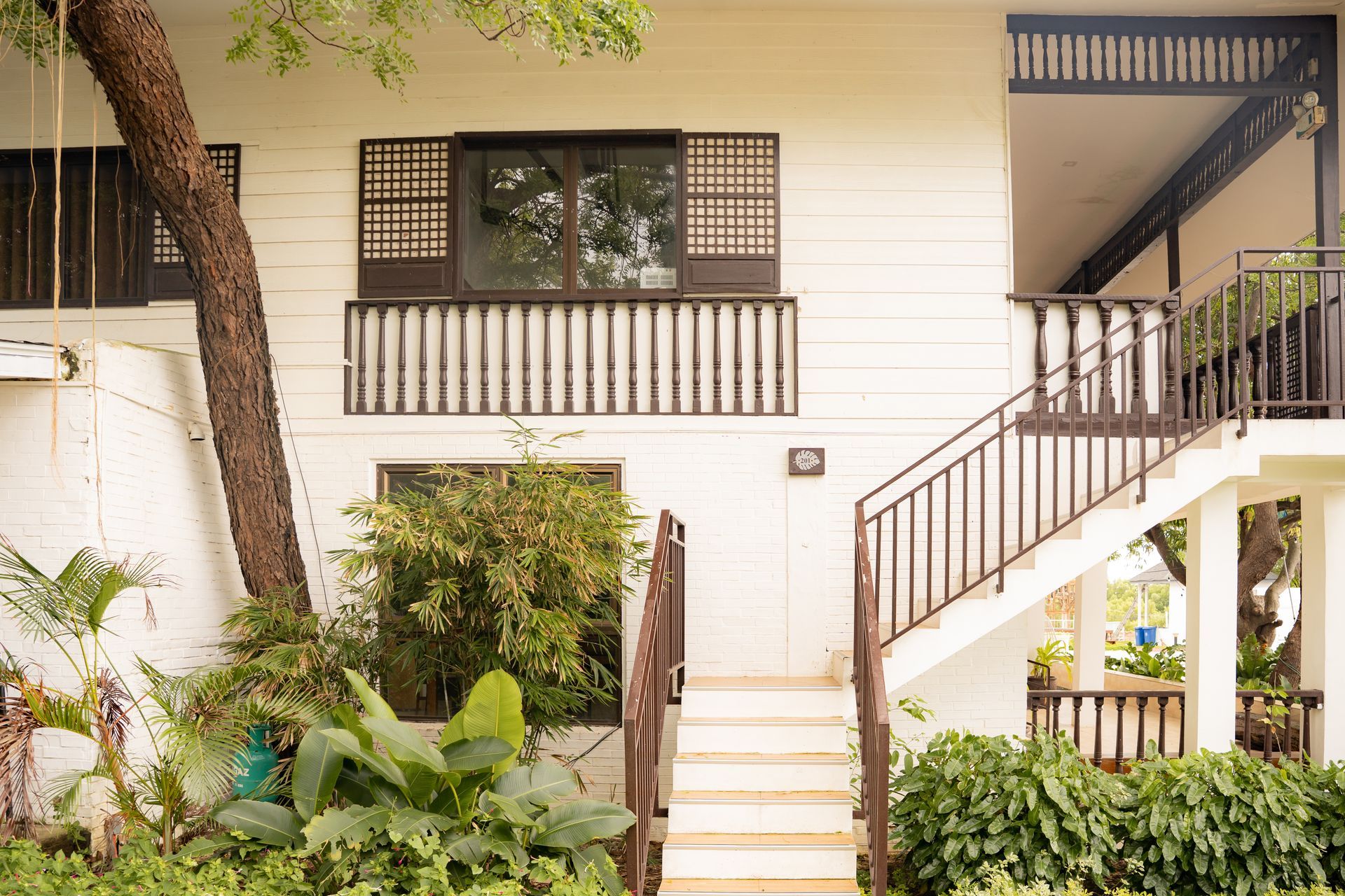 White two-story building with brown trim, stairs, and a balcony. Lush green plants surround the exterior.