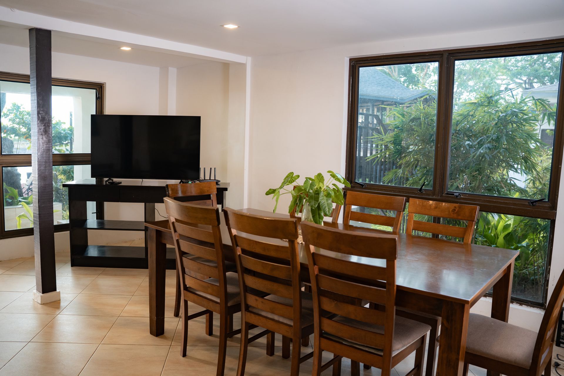Dining room with wooden table and chairs, television, and large window overlooking greenery.