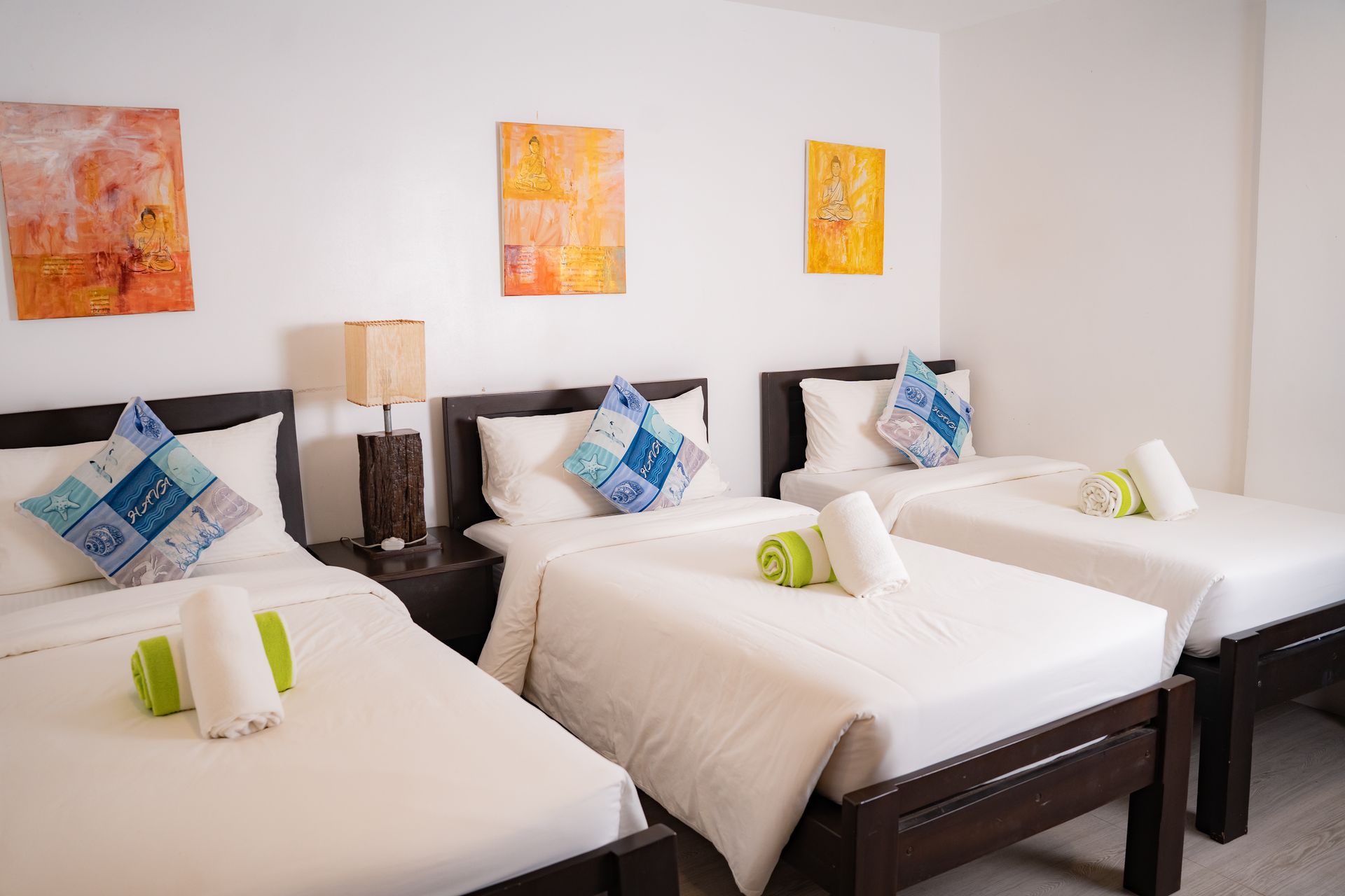 A brightly lit bedroom with three single beds, white linens, and colorful art on the wall.