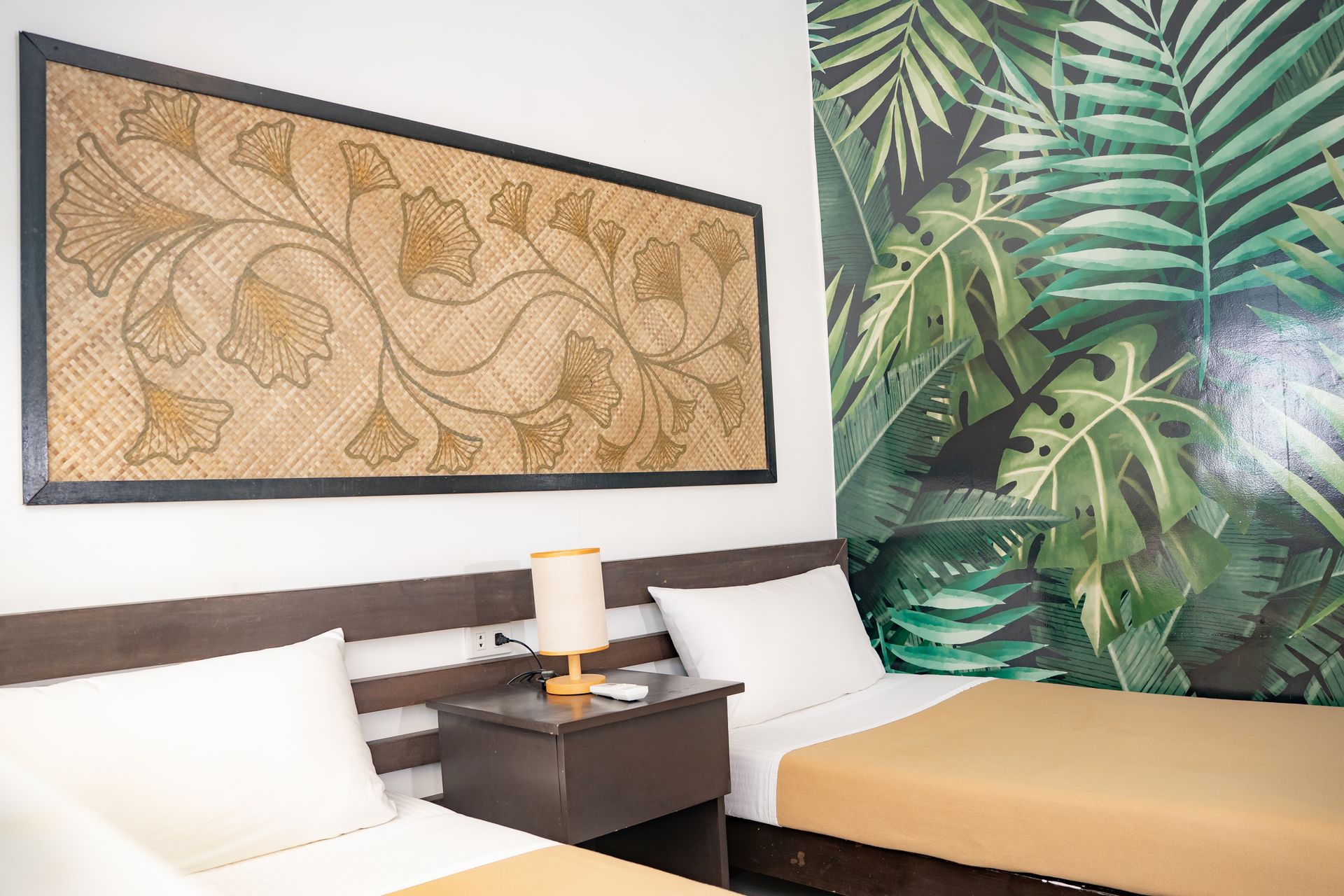 Two beds with brown sheets, nightstand, and wall art in a room with a jungle-themed accent wall.