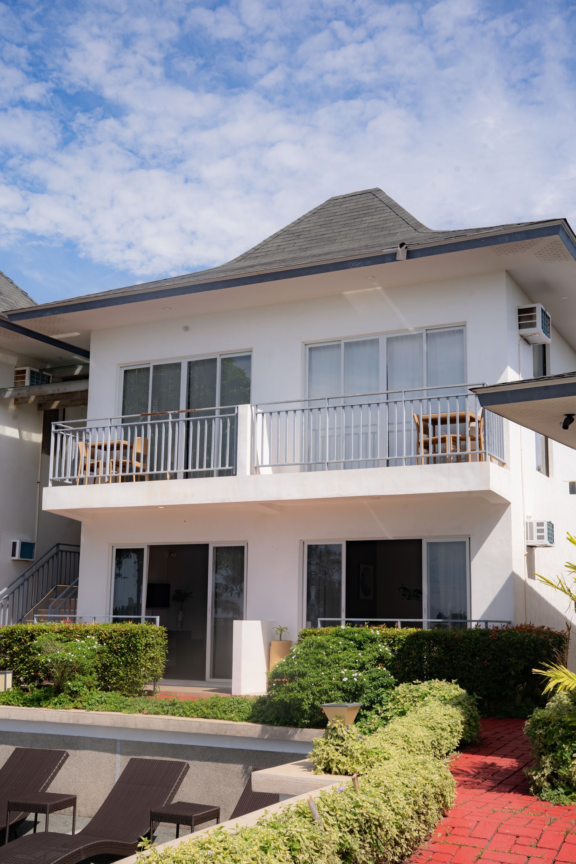 Two-story white building with a balcony, windows, and a dark roof. Lush greenery and blue sky.