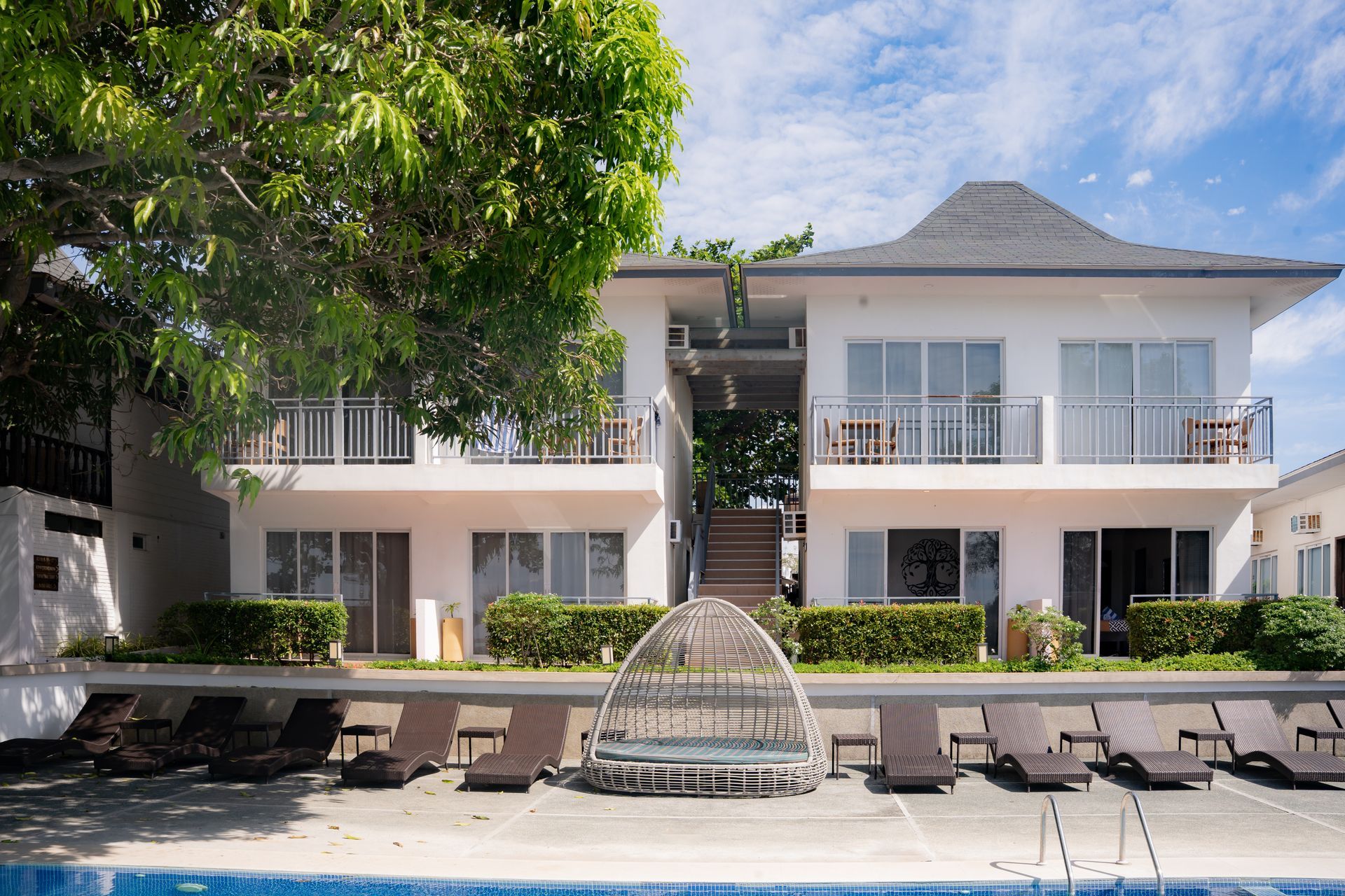 White beachfront hotel with pool, lounge chairs, and leafy trees.