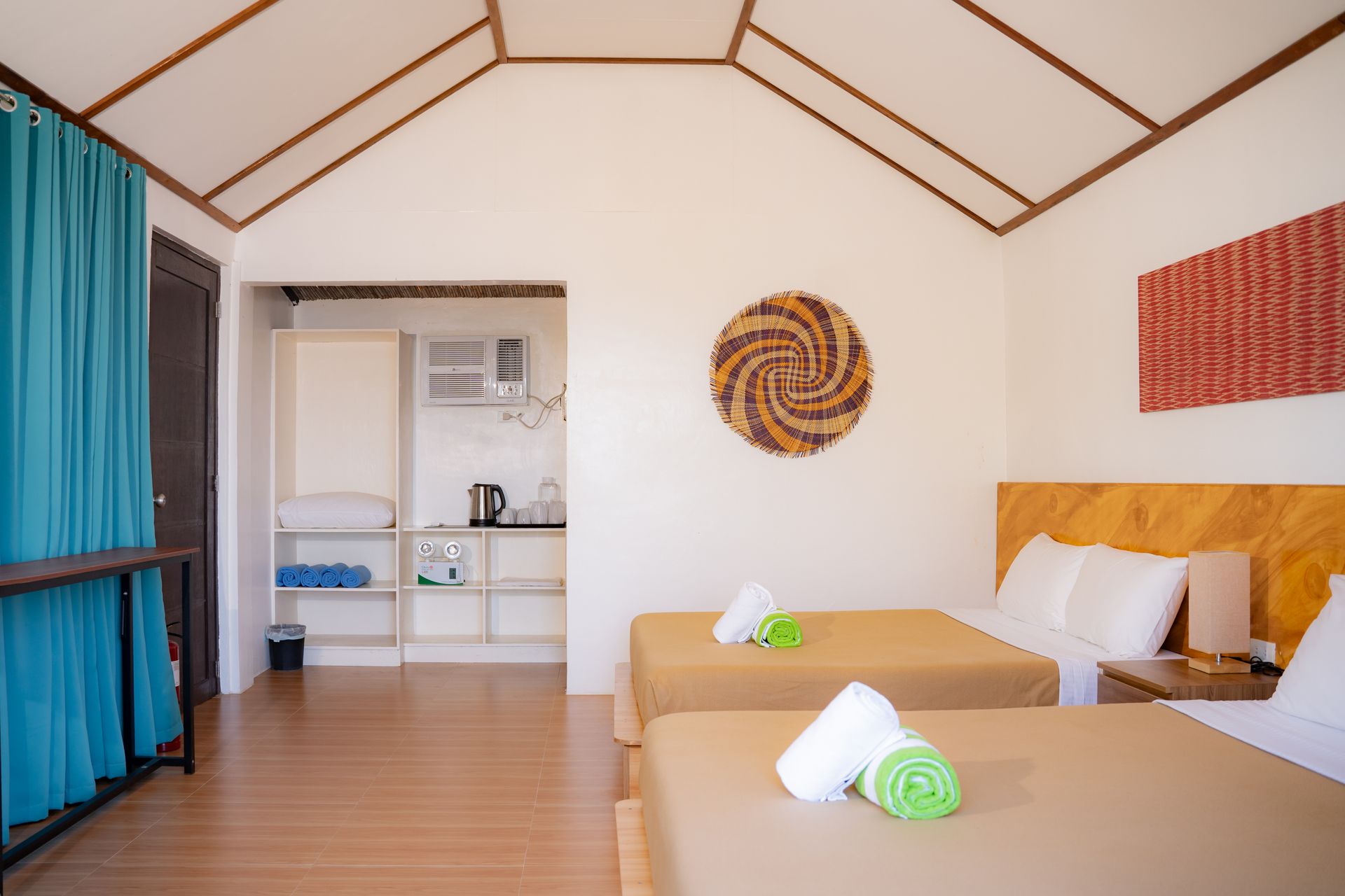 Two beds in a bright room with a high ceiling, small kitchenette, and decorative wall art.