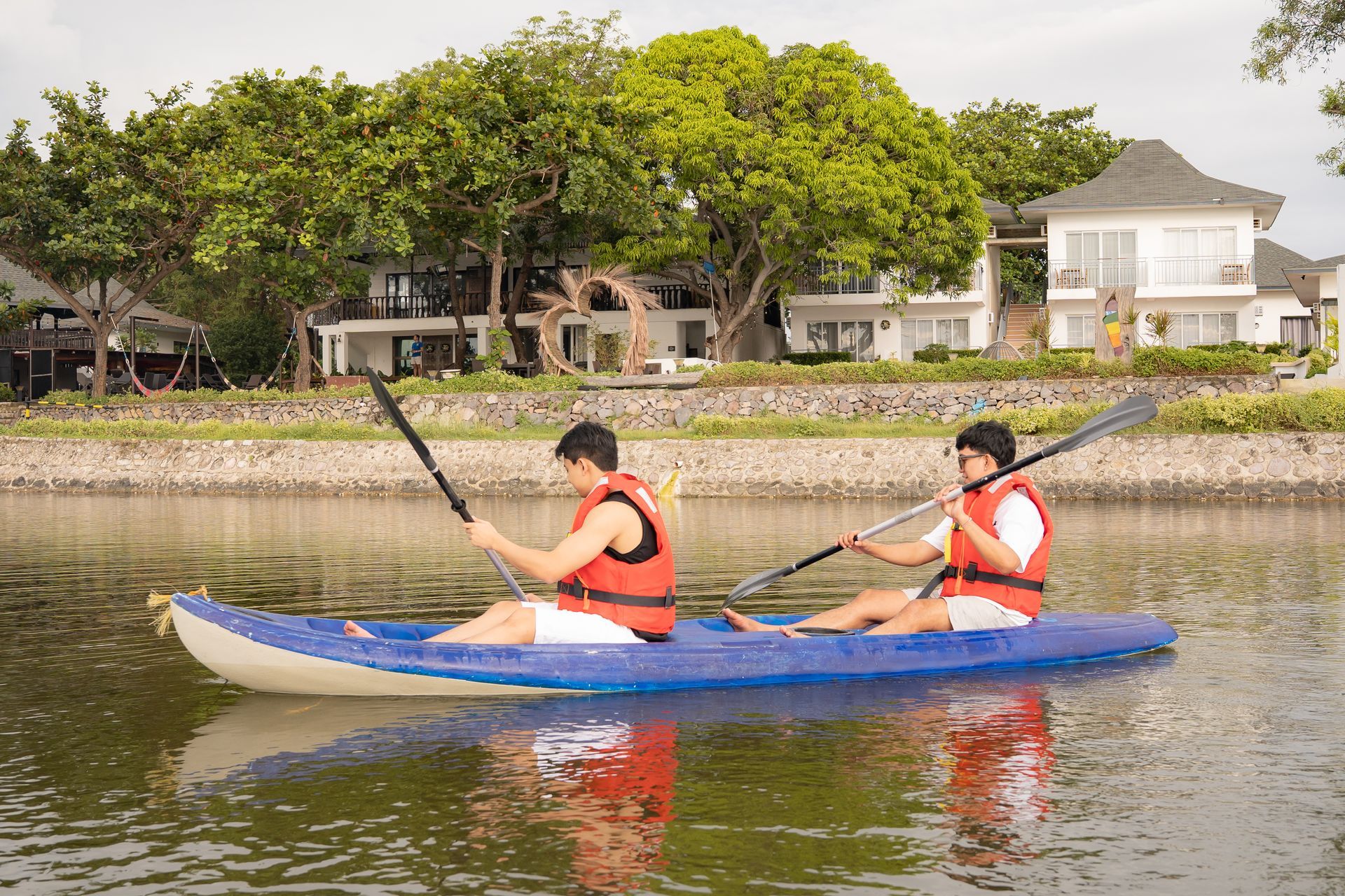 Two people kayaking in a blue kayak on calm water, wearing orange life vests. Houses and trees are in the background.
