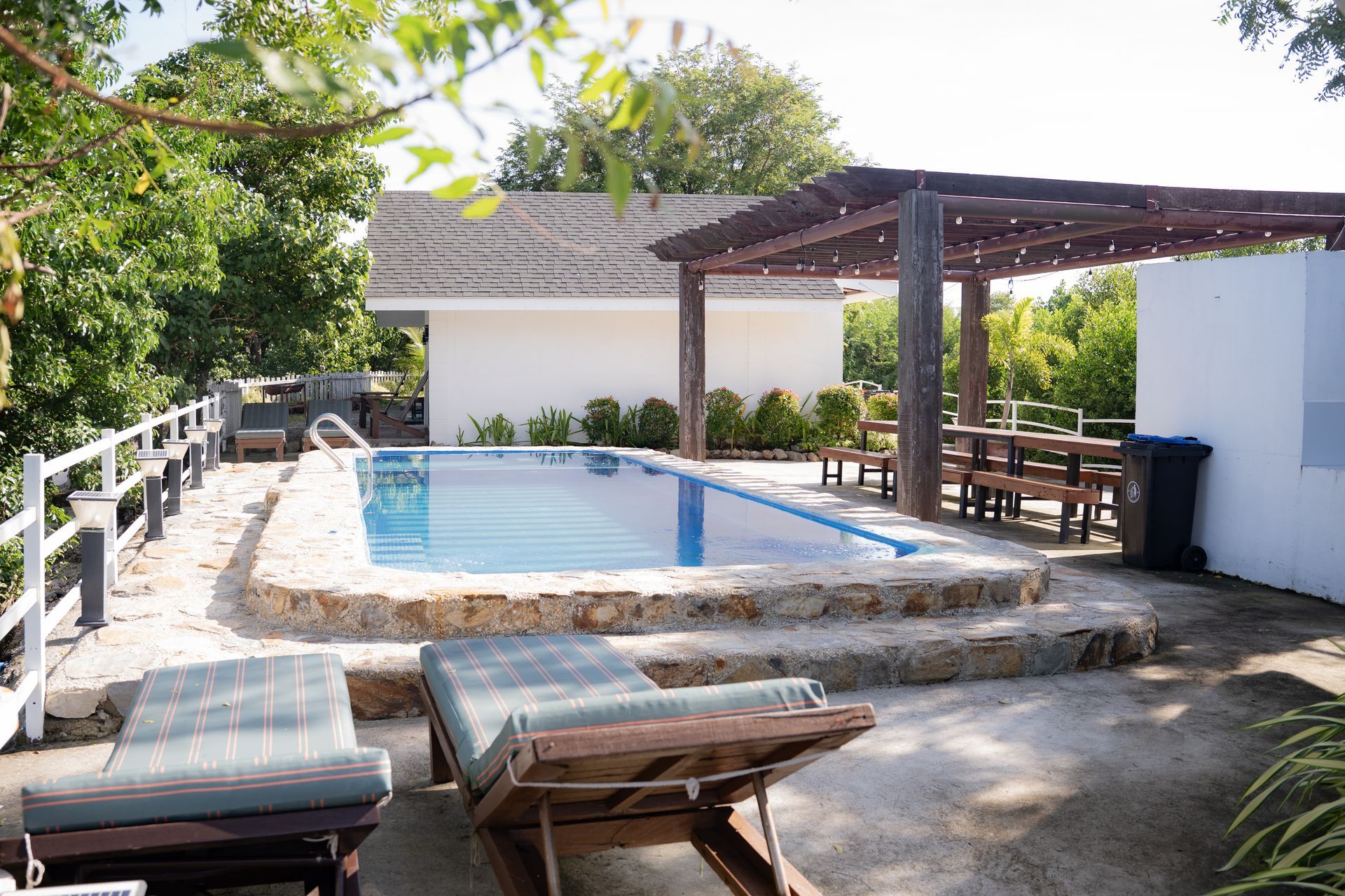 Pool with lounge chairs and shaded dining area, outside a white building.