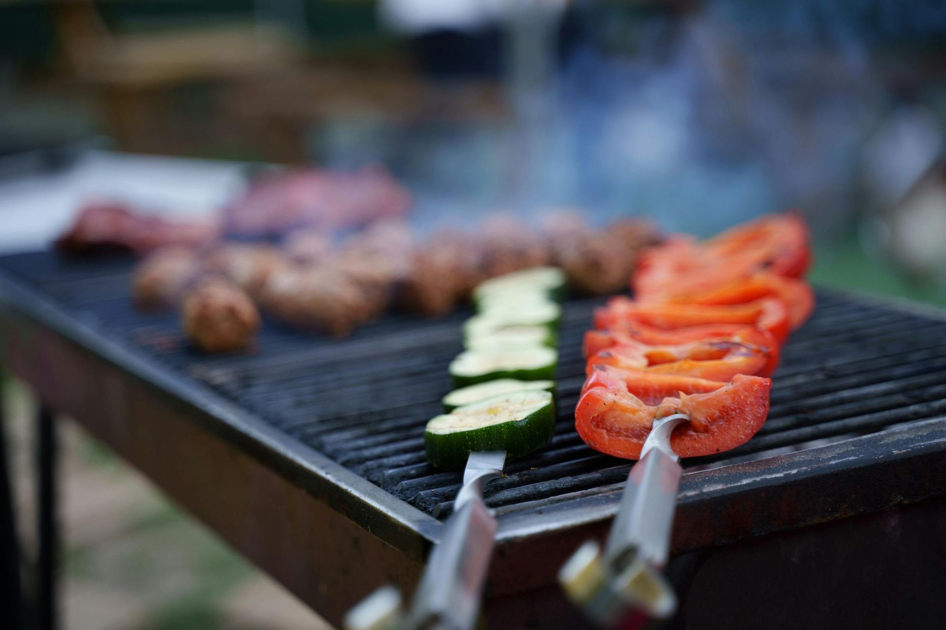 Vegetable and meat skewers grilling on a barbecue, with red peppers and zucchini in the foreground