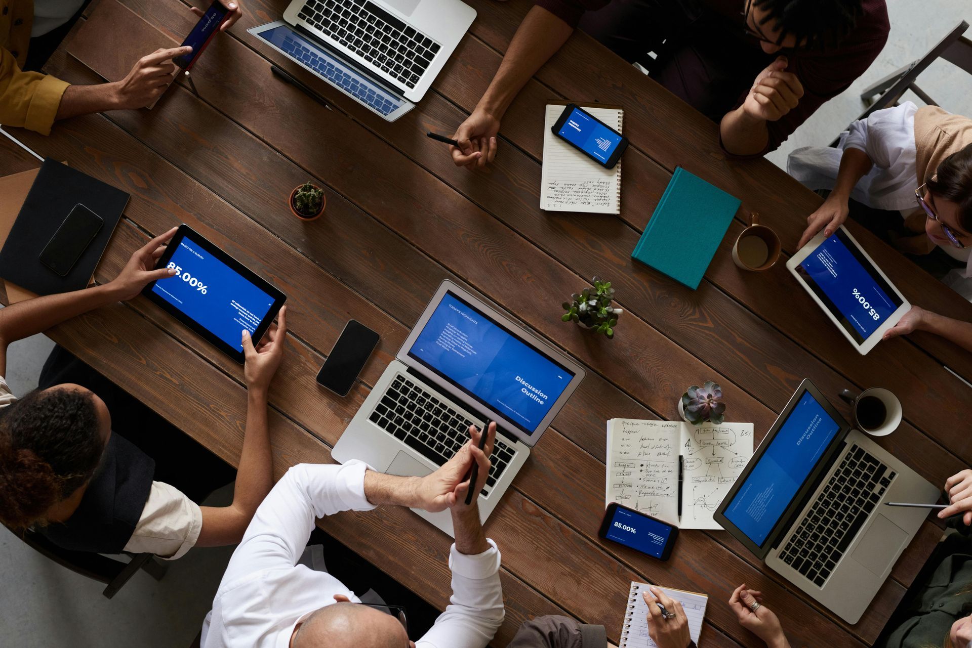 Team meeting around a wooden table with laptops, tablets, notebooks, and coffee cups.