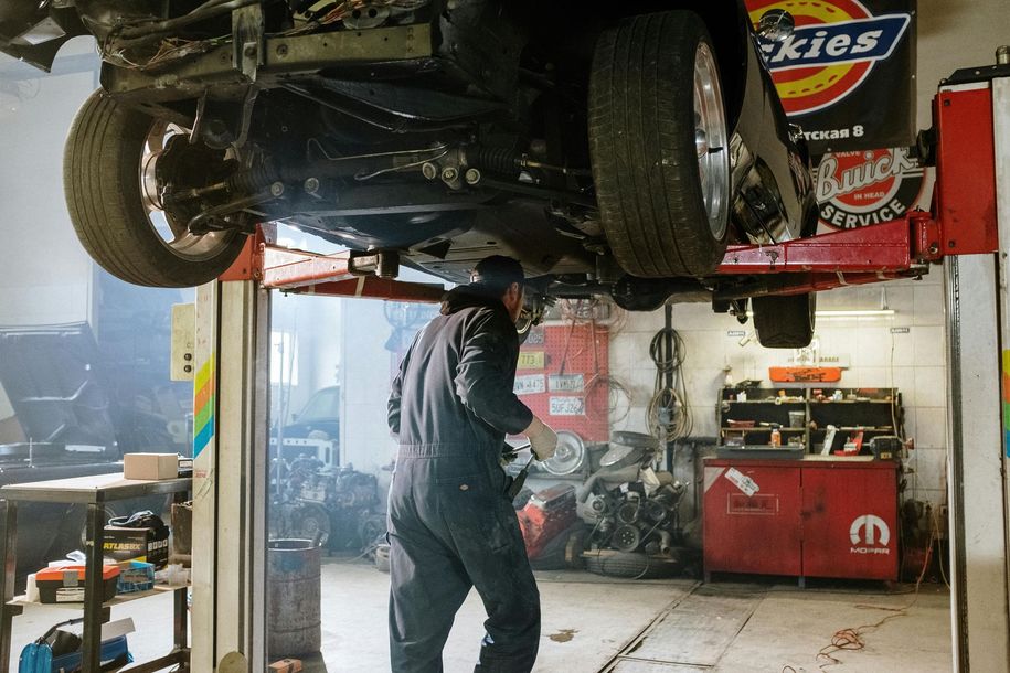 A mechanic in dark coveralls works under a car lifted on a hydraulic rack in an auto shop.