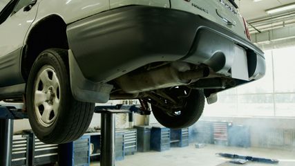 A car lifted on a hydraulic hoist inside an auto repair shop, with smoke or steam visible on the floor.
