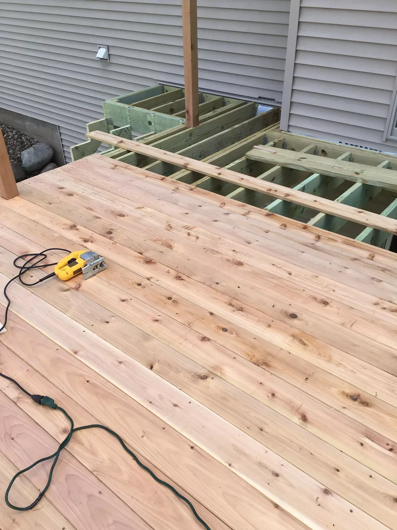 Wooden deck under construction; tools and building materials visible. Boards being laid, framework exposed, against a light siding wall.