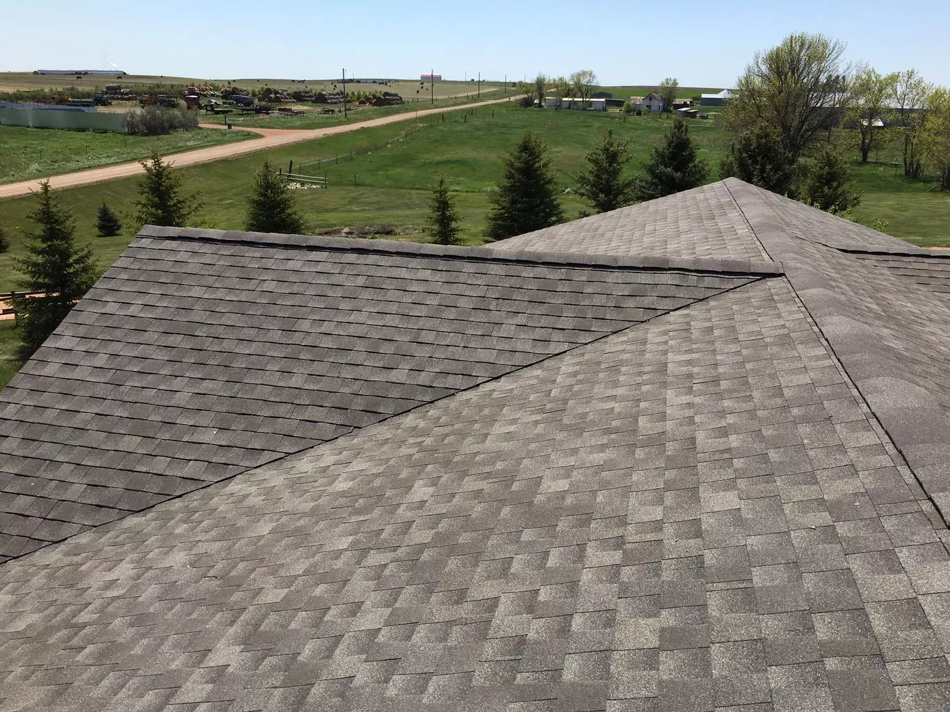 View of a roof with different shades of gray shingles. The house overlooks a green field on a sunny day.