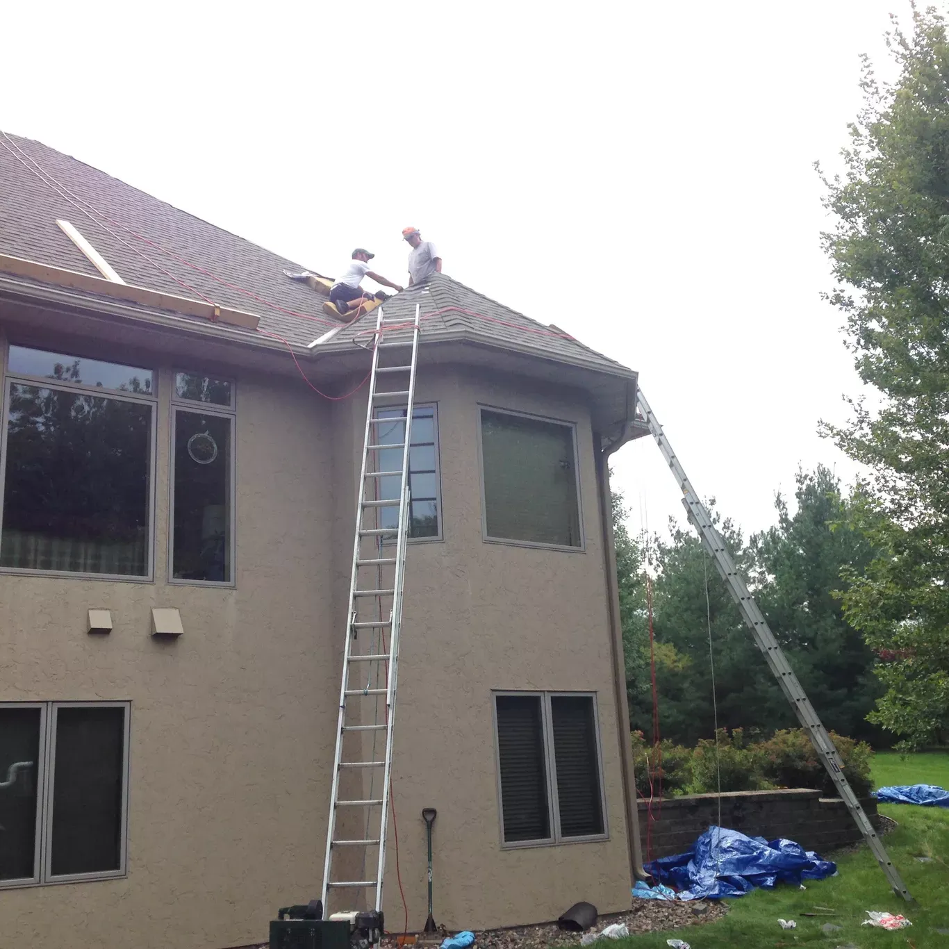 Two people on a roof, using two ladders. One person is working on the roof's edge. The house is beige with brown roofing.