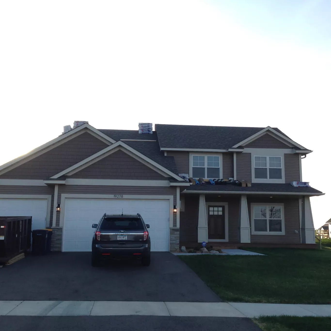 A two-story house with a dark SUV in the driveway. The house has a brown exterior and white garage doors.