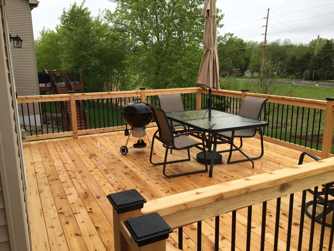 Wooden deck with a table, chairs, grill, and umbrella, next to a house. Black railing and green trees in the background.